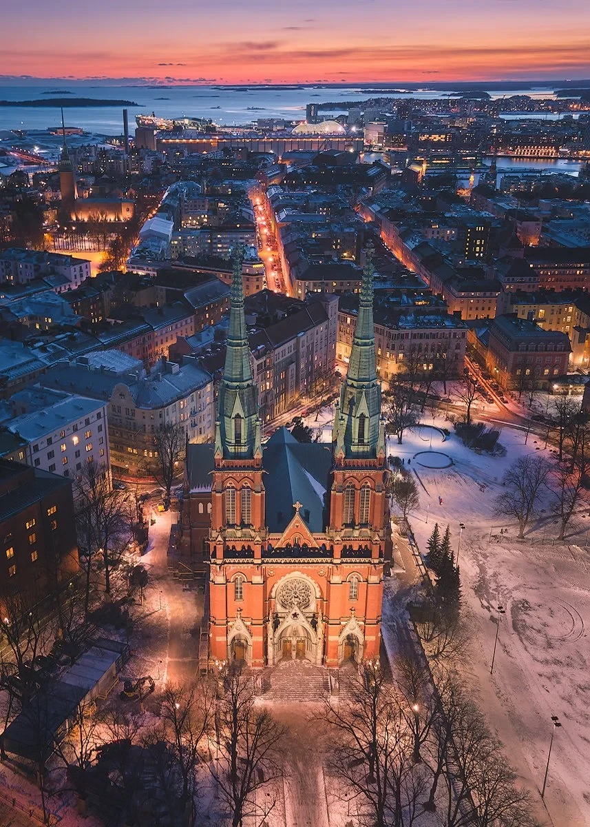Aerial view of Helsinki cityscape at sunset with a prominent historic church, snow-covered streets, and illuminated city lights.