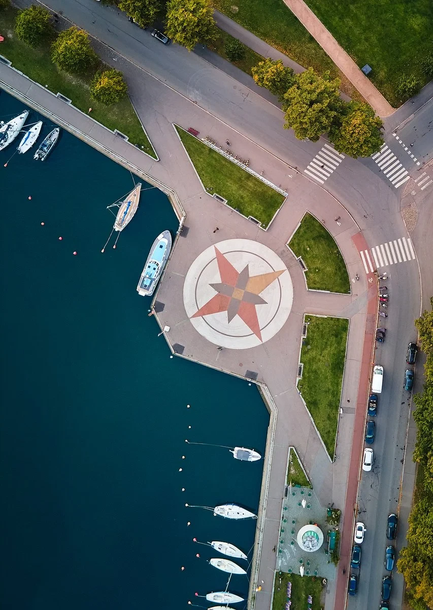 Aerial view of a waterfront area with small boats docked along a curved quay. A large compass rose design is on the ground near the water. Roads border the area, with cars parked along the street and trees providing some greenery.