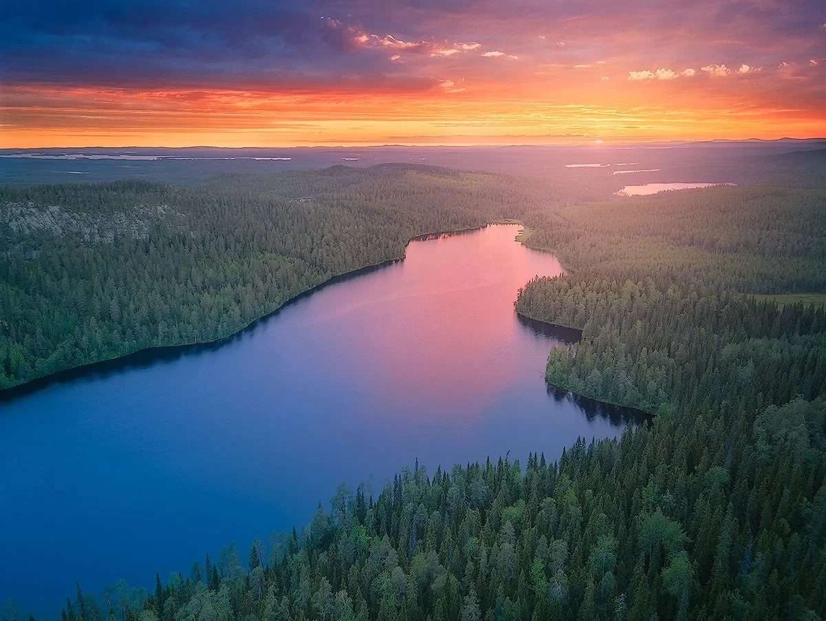 Aerial view of a winding river surrounded by dense forest at sunset, with a colorful sky filled with orange and purple hues.