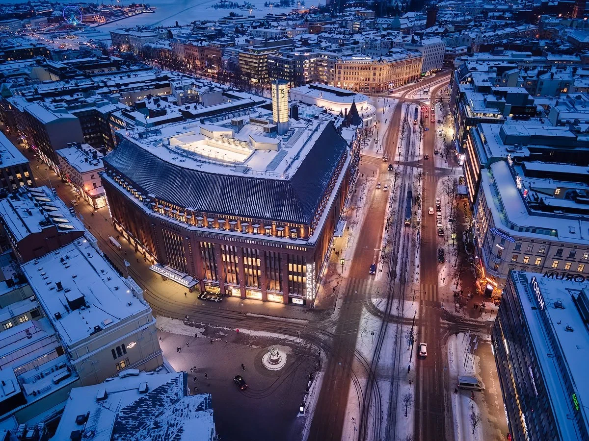Aerial view of a snowy cityscape with buildings and roads illuminated at dusk. A large, illuminated building with a distinct architectural design is prominent in the center. Streets are lined with vehicles and city lights, and there's a visible snow