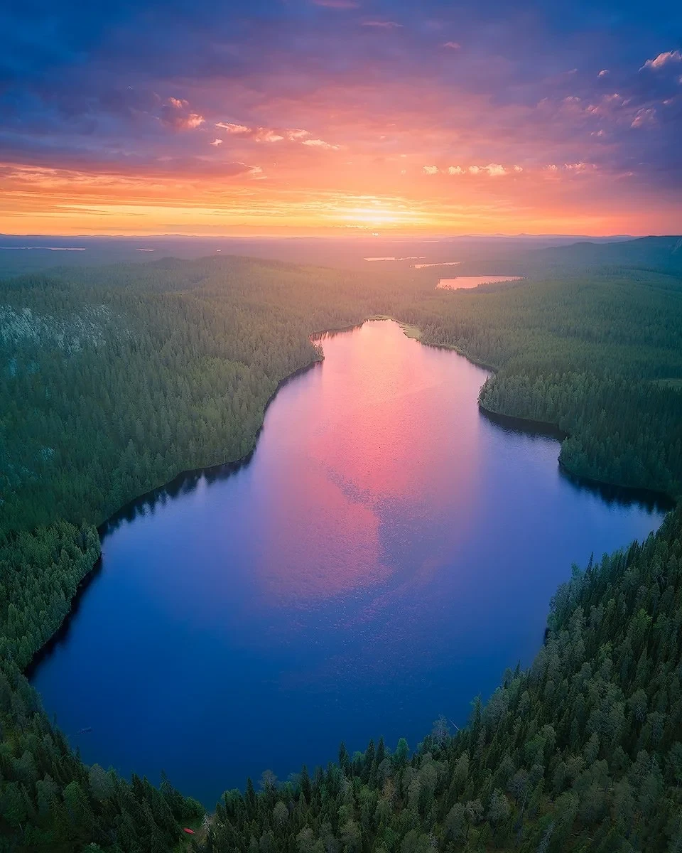Aerial view of a serene lake surrounded by dense forest, with a vibrant sunset casting a pink and orange glow on the water and sky.