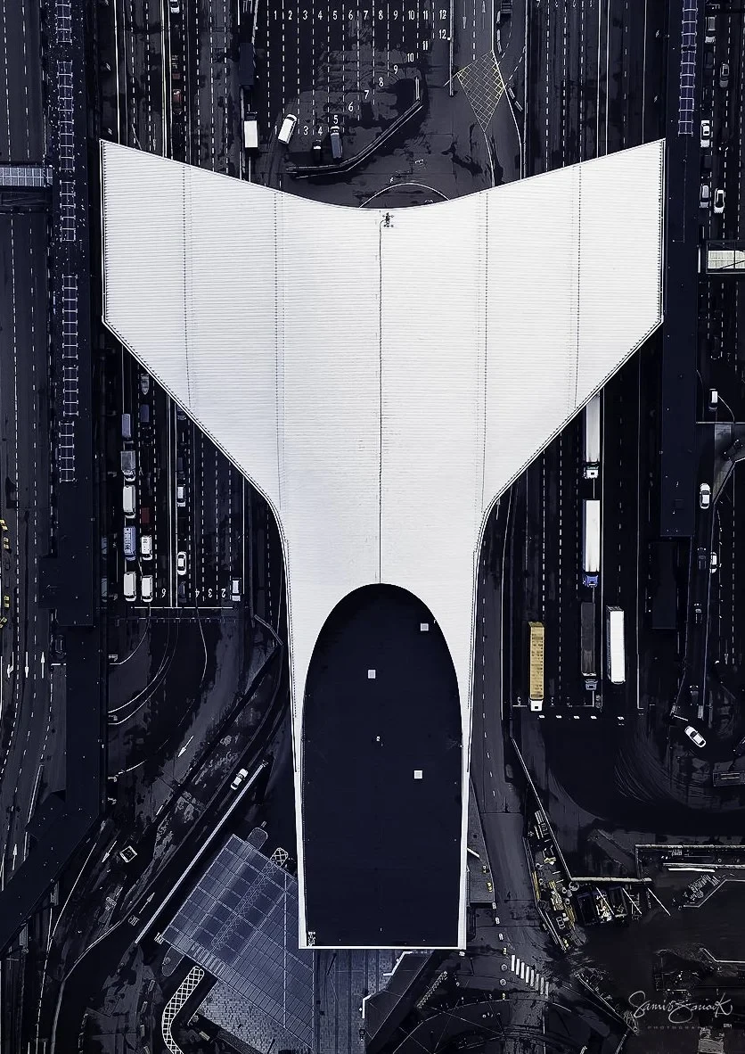 Aerial view of a large, white-roofed building with curved edges surrounded by multiple lanes of traffic, vehicles visible on the roads.