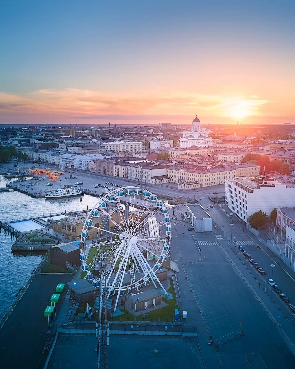 Aerial view of Helsinki with a Ferris wheel in the foreground, overlooking the cityscape at sunset. Notable buildings and waterfront visible.