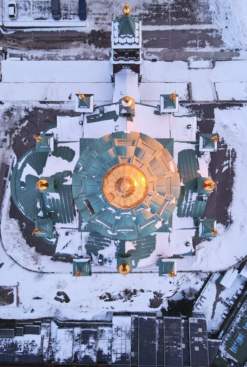 Aerial view of a snow-covered building with a green roof and gold domes, surrounded by snowy streets and parked cars.