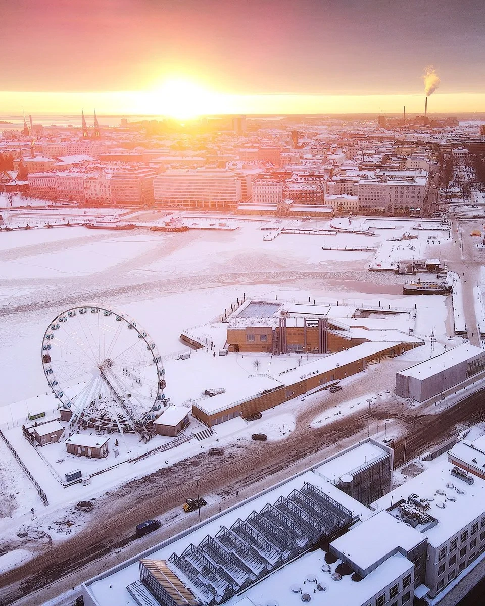 Aerial view of a cityscape during winter with a Ferris wheel, snow-covered buildings, and a frozen harbor. The sun is setting on the horizon, casting a warm glow over the cold scene. Smoke rises from a distant chimney, and streets are lightly dusted